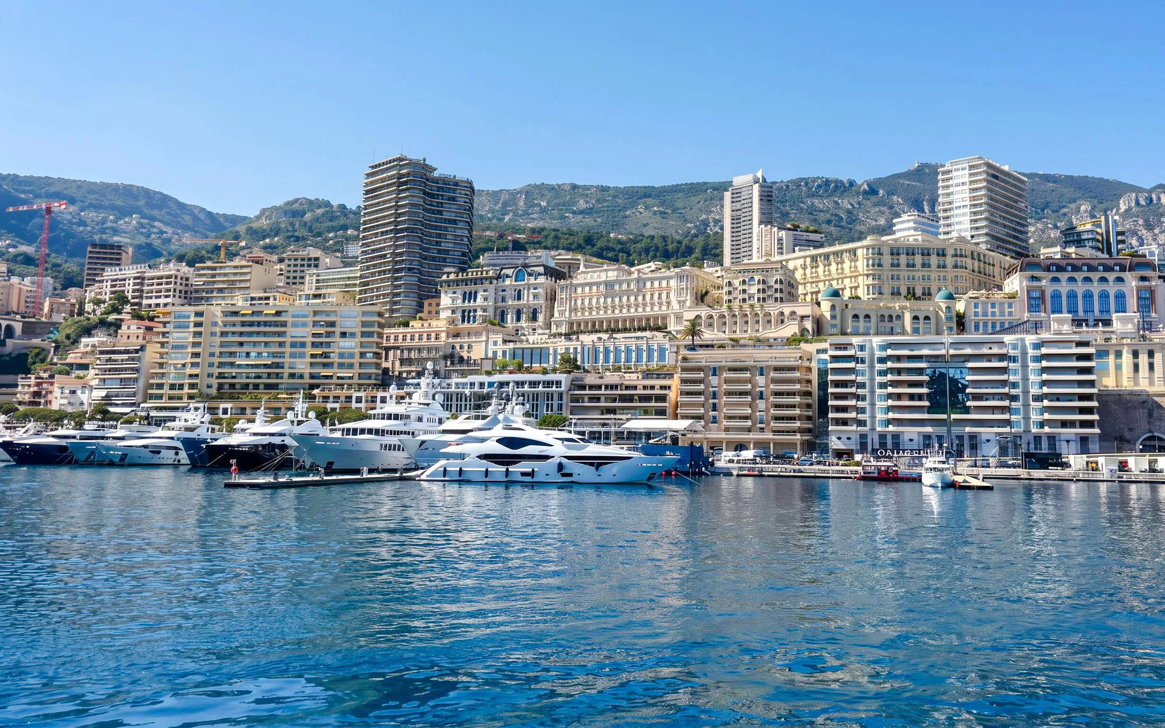 Yachts docked in Monaco harbor with cityscape and hills in the background.
