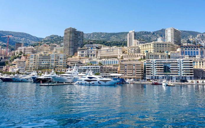Yachts docked in Monaco harbor with cityscape and hills in the background.