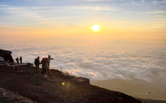 Climbers on Mt. Fuji summit at sunrise, overlooking a sea of clouds.