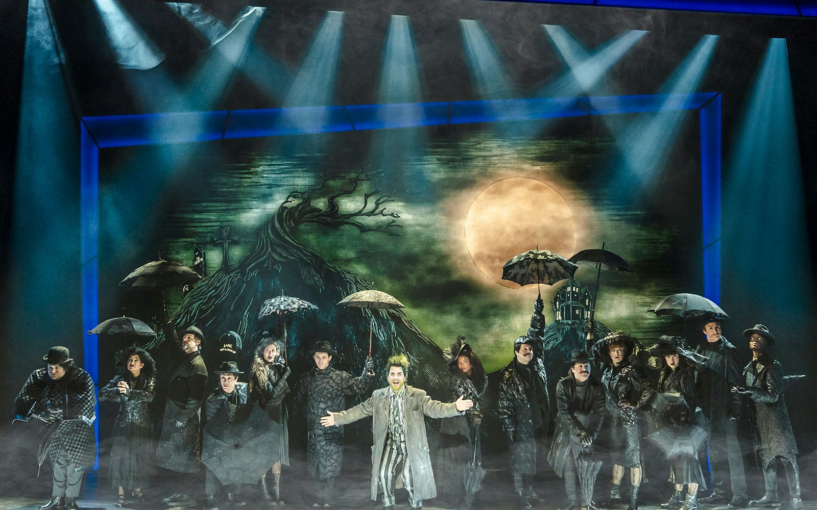 Performer on stage at Beetlejuice Broadway show with cast under umbrellas.