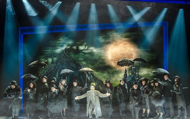 Performer on stage at Beetlejuice Broadway show with cast under umbrellas.