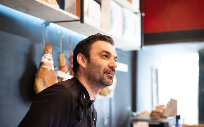 Man in a deli shop in Verona with hanging cured meats in the background.