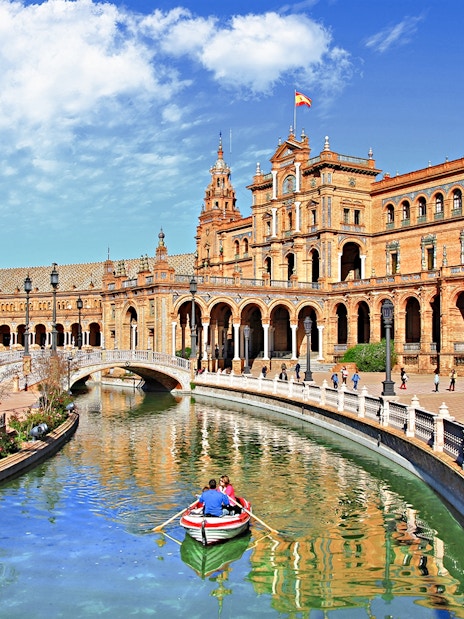 Rowboat on canal at Plaza de España, Seville, with historic building in background.