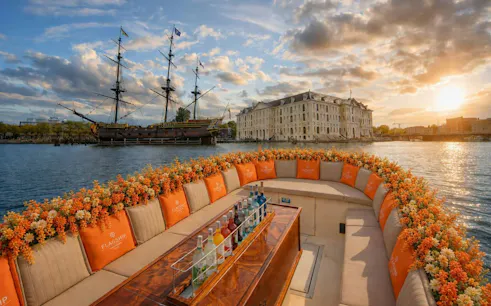 Luxury boat with floral decor cruising Amsterdam canal at sunset, near historic ship and building.