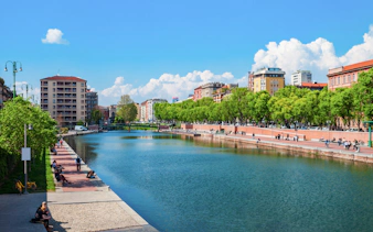 Navigli canals in Milan with people walking along the tree-lined promenade.
