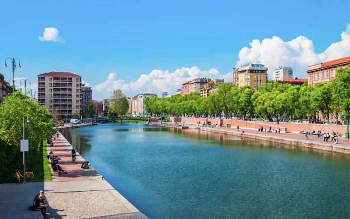Navigli canals in Milan with people walking along the tree-lined promenade.
