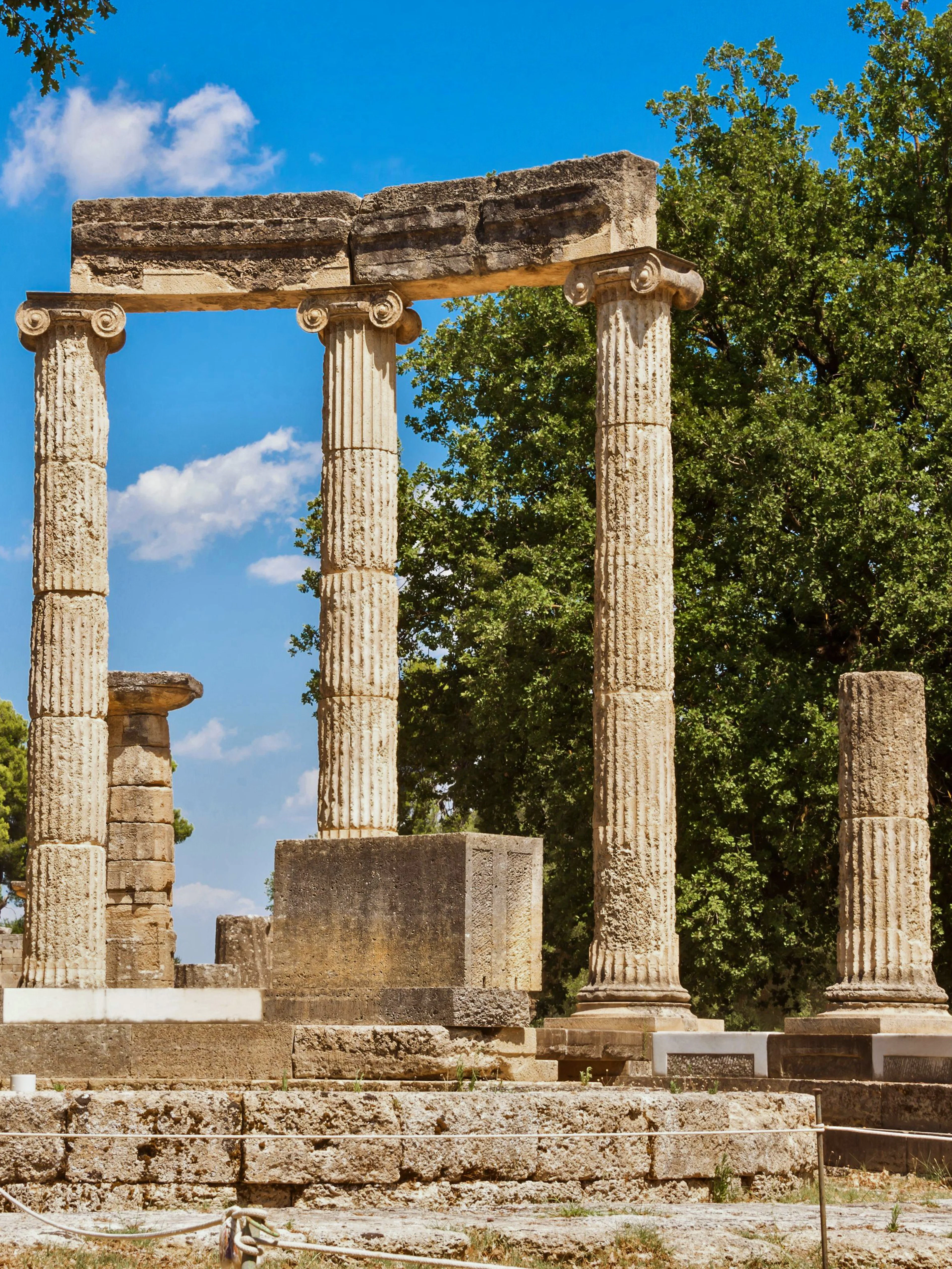 Ancient Greek columns at Olympia ruins, Greece, surrounded by trees.