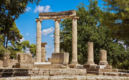 Ancient Greek columns at Olympia ruins, Greece, surrounded by trees.