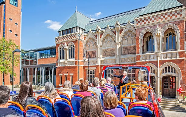 Open-top bus tour in Stratford-upon-Avon passing by historic building with guide speaking to passengers.