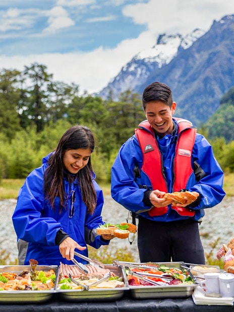 People enjoying a picnic with food trays in Dart River Funyaks, New Zealand.