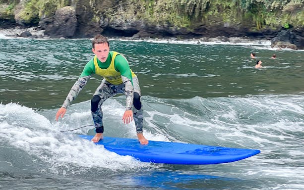 Surfer riding a wave during a family surf lesson near rocky coastline.