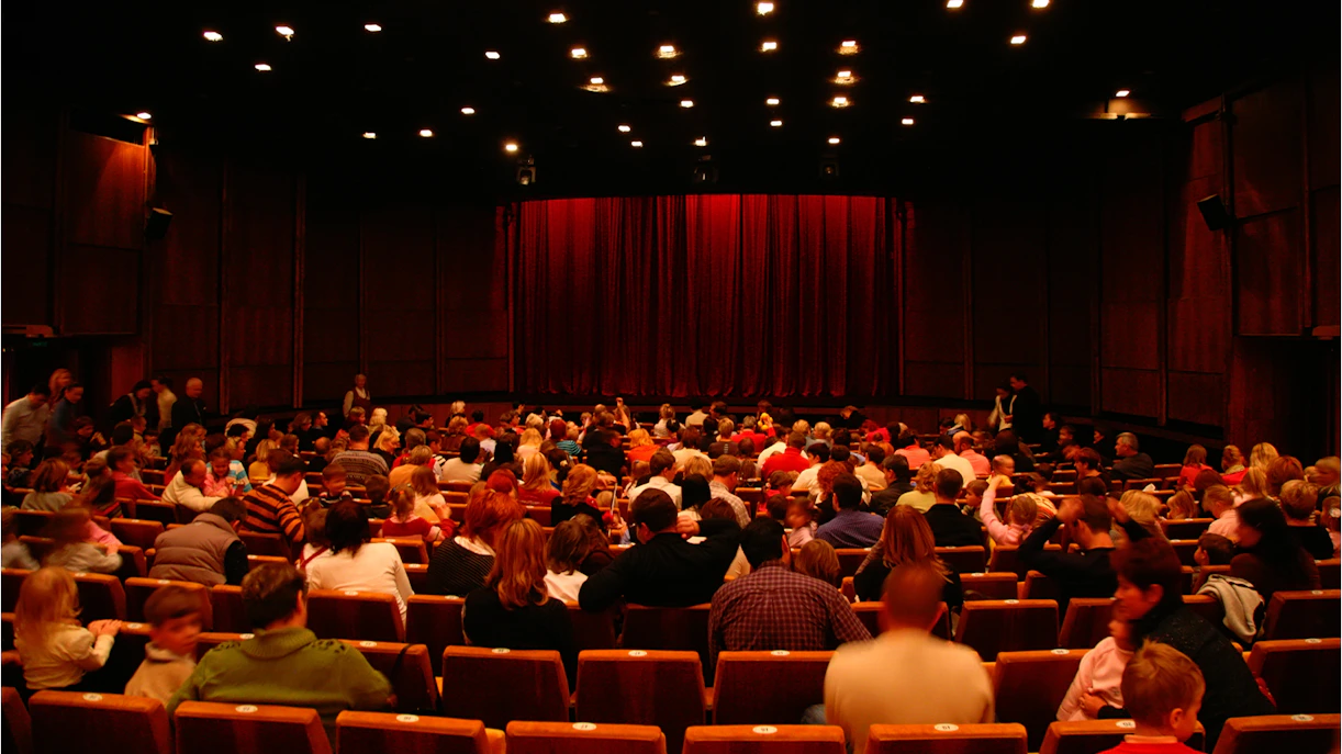 Audience seated in a West End theatre with red curtains on stage.