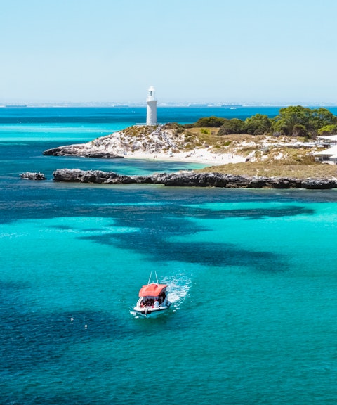 Small red boat in turquoise water near a lighthouse and island.