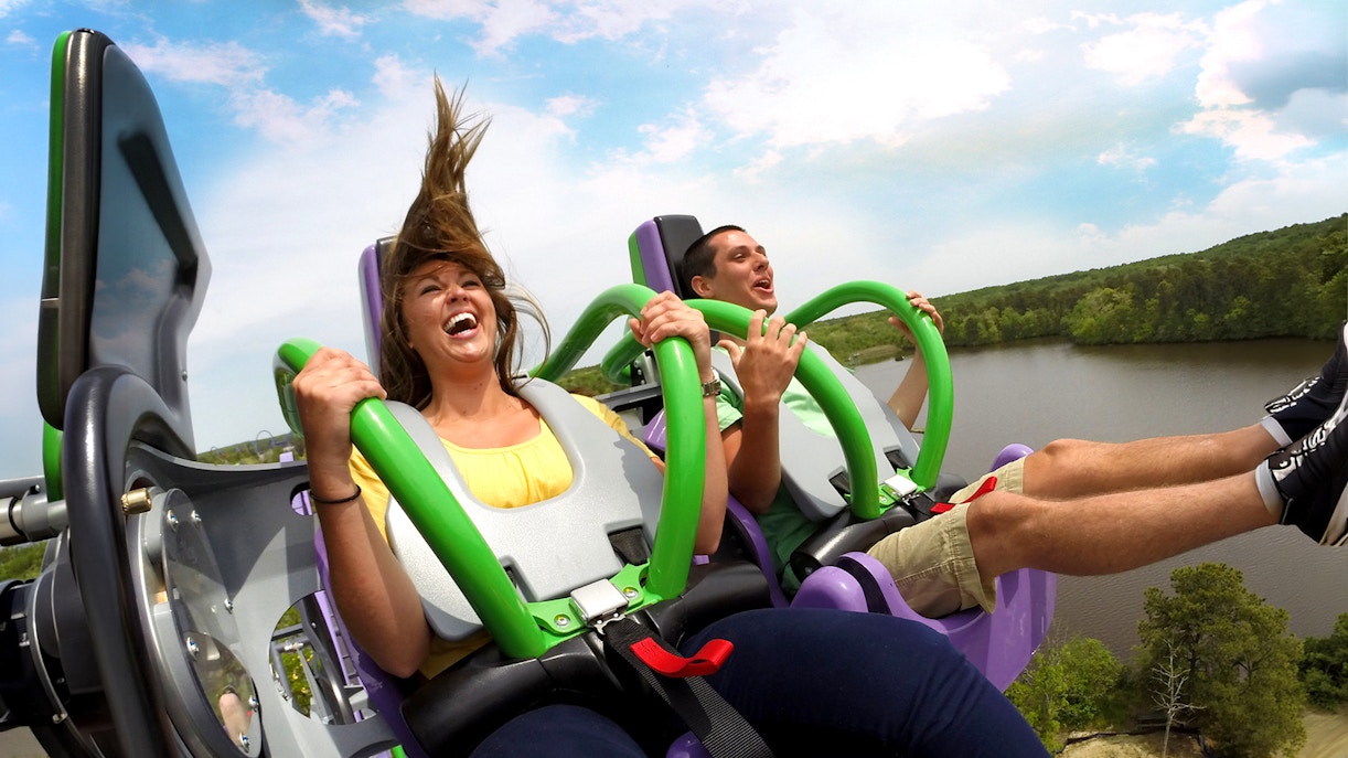 Riders enjoying THE JOKER™ roller coaster at Six Flags Great Adventure with a lake view.