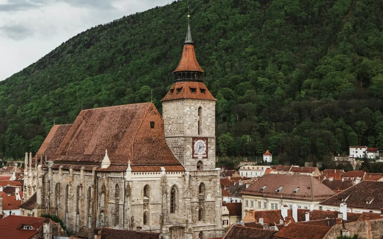 Medieval church with clock tower in Brasov, surrounded by forested hills.
