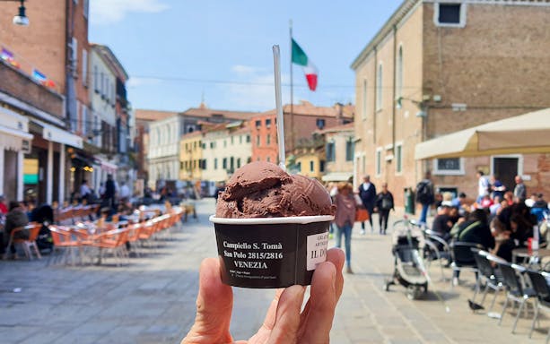 Chocolate gelato held up in a Venetian street with outdoor cafes and Italian flag in the background.