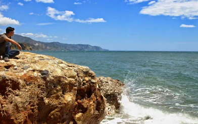 Person sitting on rocky coastline with view of Nerja, Spain.
