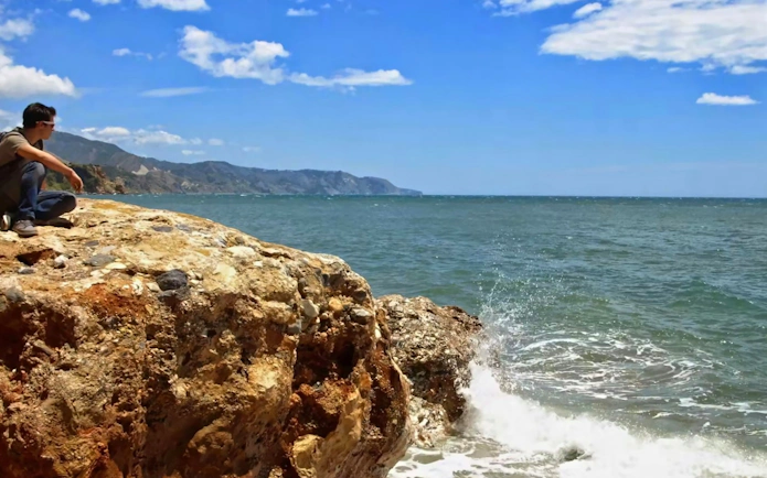 Person sitting on rocky coastline with view of Nerja, Spain.