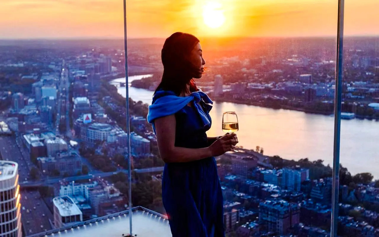 Person enjoying sunset view over Boston skyline with a glass of wine.