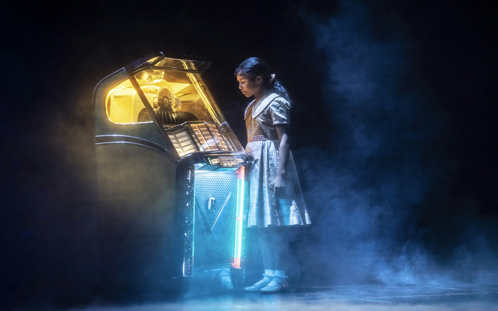 Young girl standing by a glowing jukebox in a scene from The Drifters Girl musical.