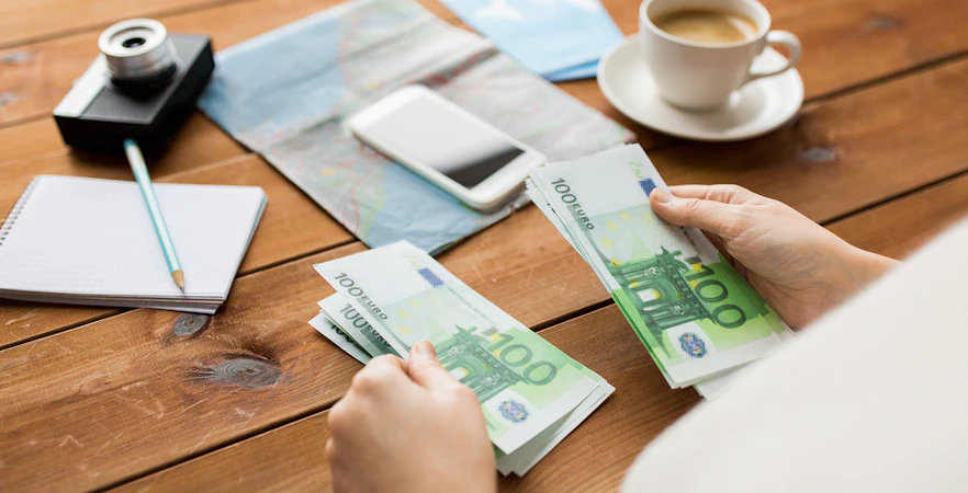 Hands counting 100 euro bills on a table with a map, camera, and coffee.