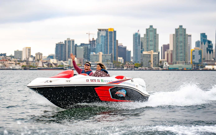 Speed boat with two people on San Diego Bay, city skyline in background.