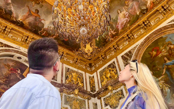 Visitors admiring ornate ceiling and chandelier in Versailles palace.
