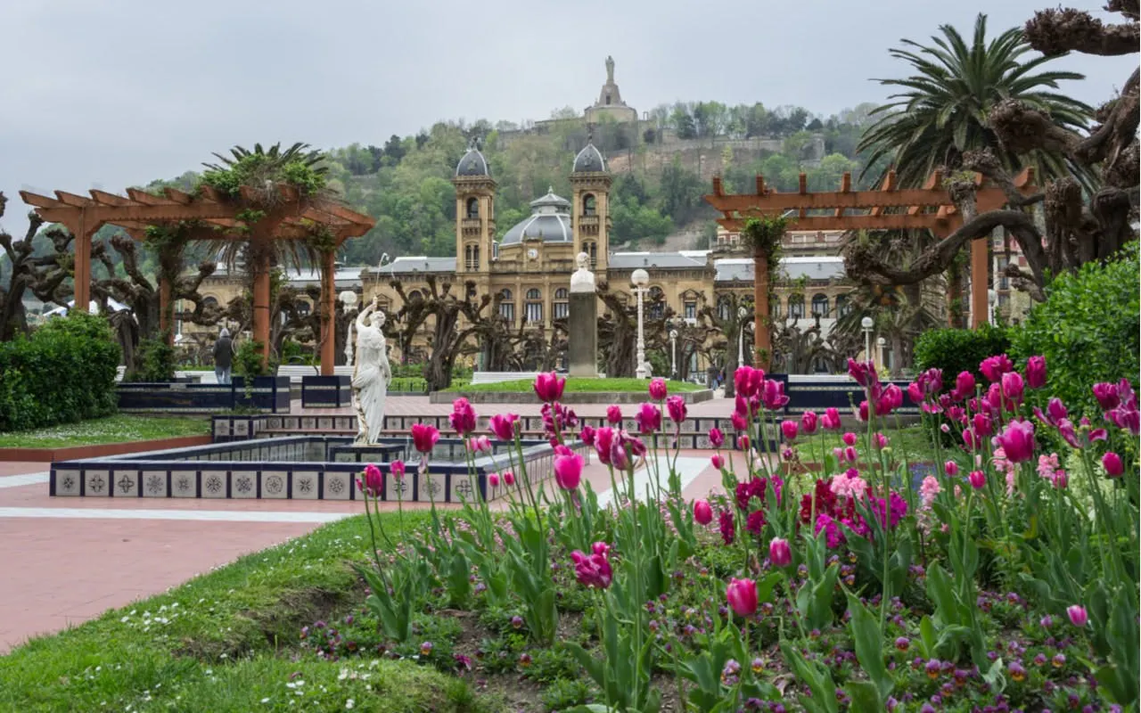 San Sebastian city garden with pink tulips, historic building, and distant hill statue.