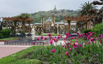 San Sebastian city garden with pink tulips, historic building, and distant hill statue.