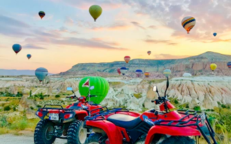 ATVs parked with hot air balloons over Cappadocia landscape at sunset.