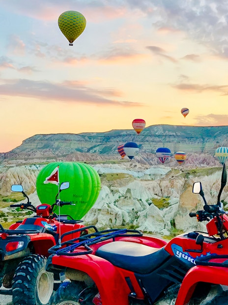 ATVs parked with hot air balloons over Cappadocia landscape at sunset.