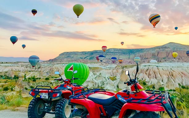 ATVs parked with hot air balloons over Cappadocia landscape at sunset.