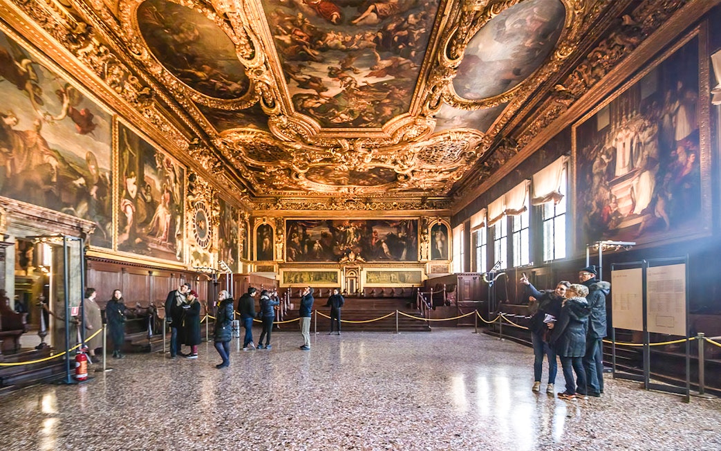 Visitors inside the ornate hall of Doge's Palace, Venice, with detailed ceiling frescoes.
