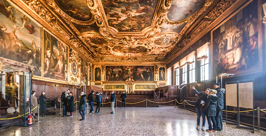 Visitors inside the ornate hall of Doge's Palace, Venice, with detailed ceiling frescoes.
