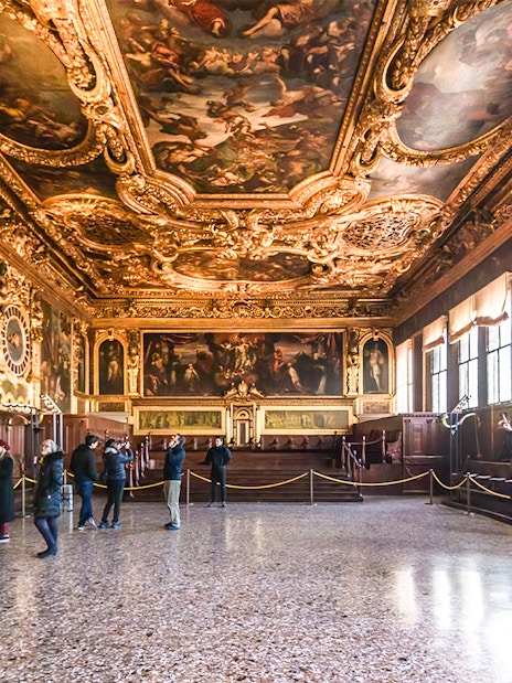 Visitors inside the ornate hall of Doge's Palace, Venice, with detailed ceiling frescoes.