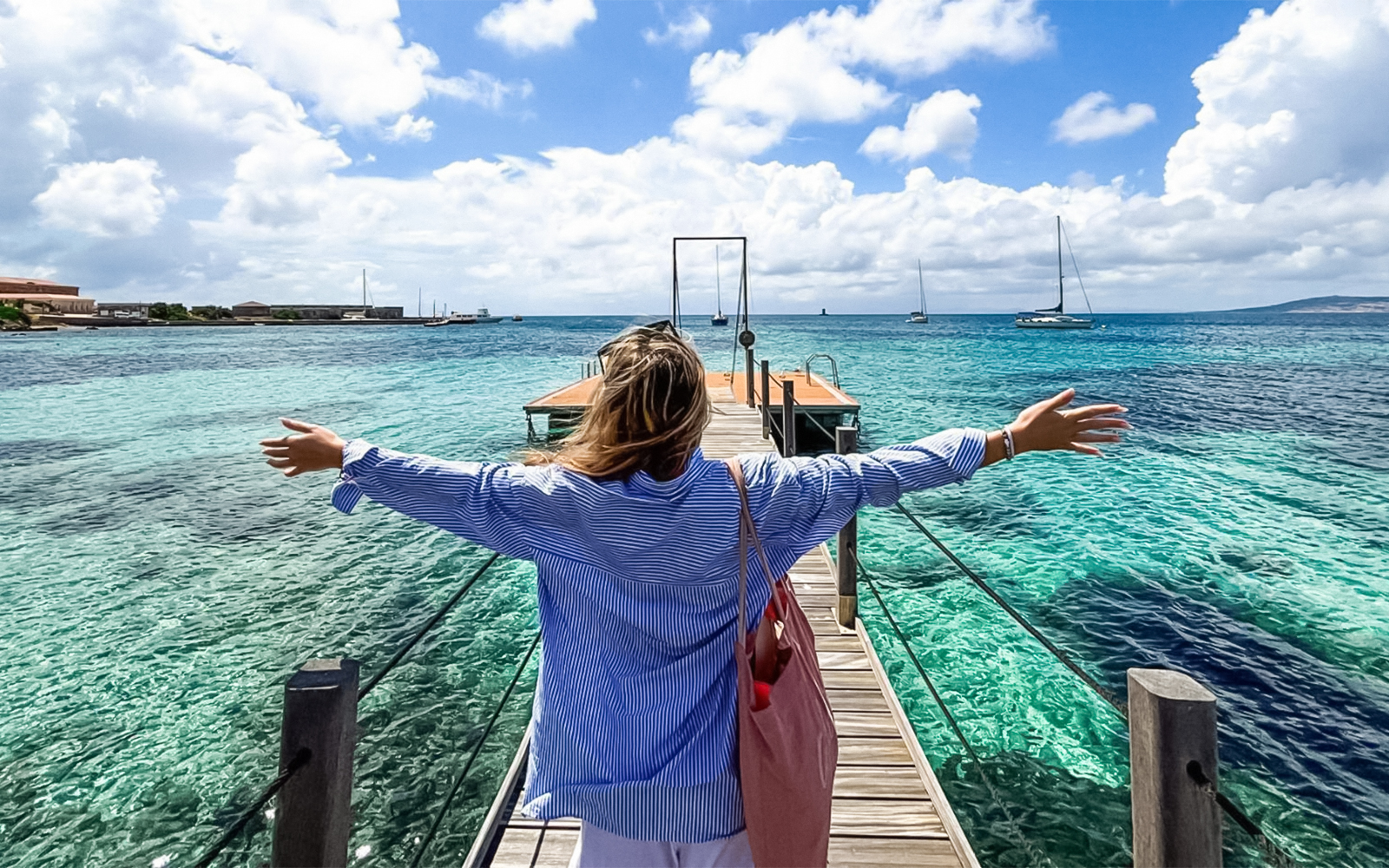 Person on a dock overlooking turquoise waters with sailboats near Asinara Island.