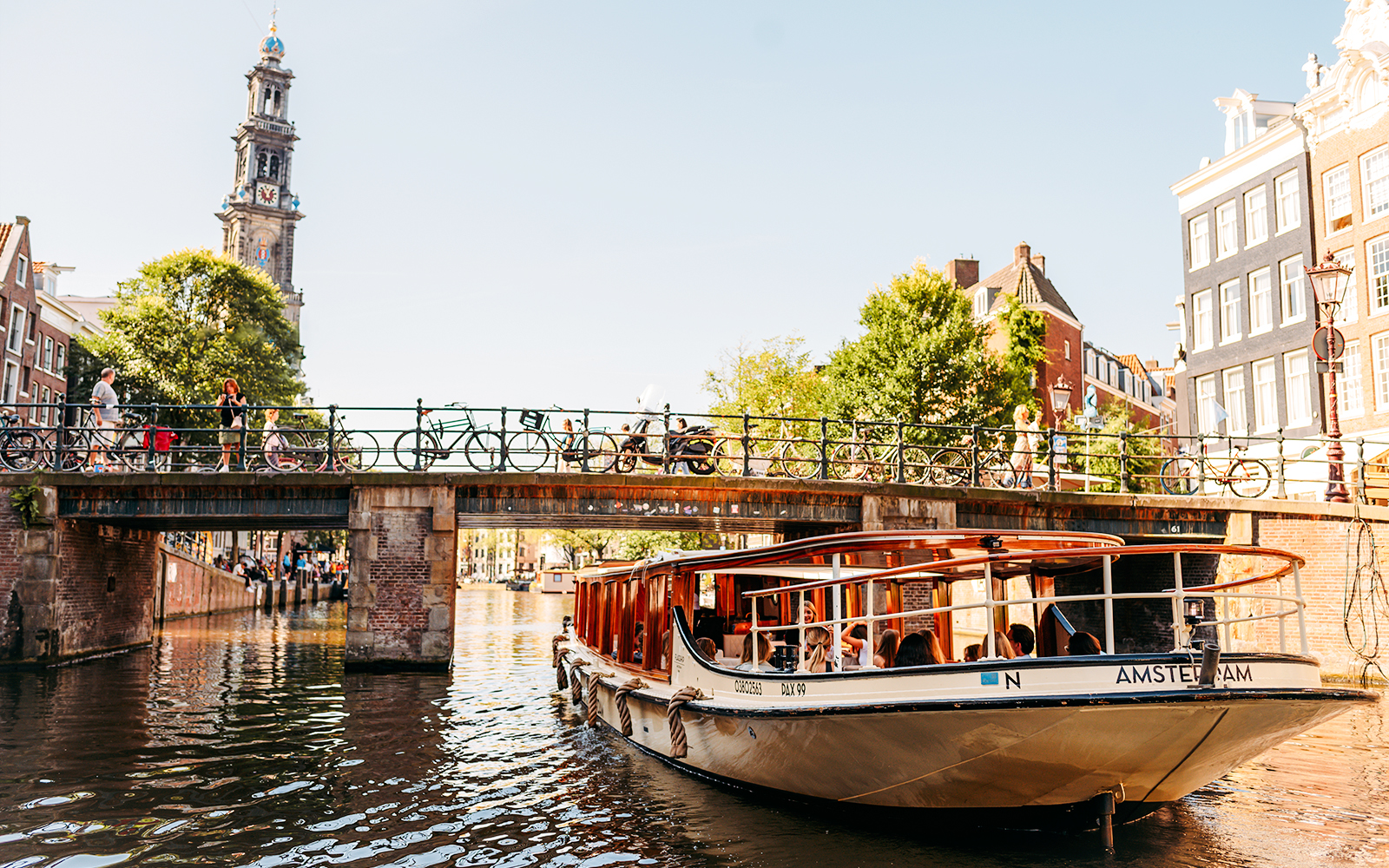 Amsterdam canal cruise boat passing under a bridge with Westerkerk tower in the background.