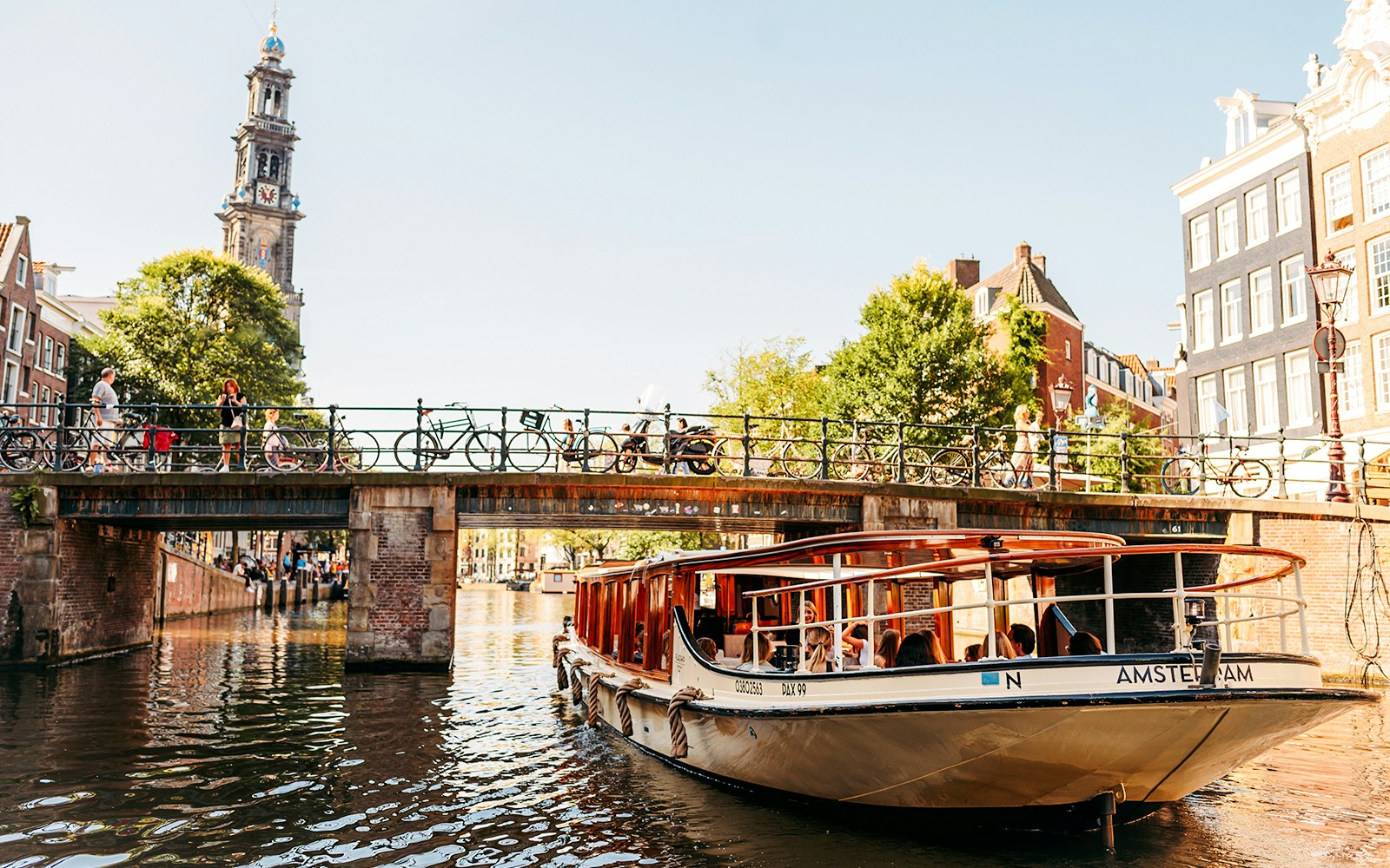 Amsterdam canal cruise boat passing under a bridge with Westerkerk tower in the background.