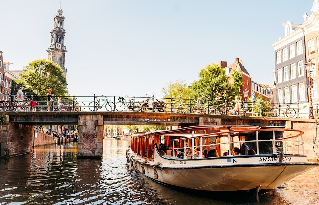 Amsterdam canal cruise boat passing under a bridge with Westerkerk tower in the background.