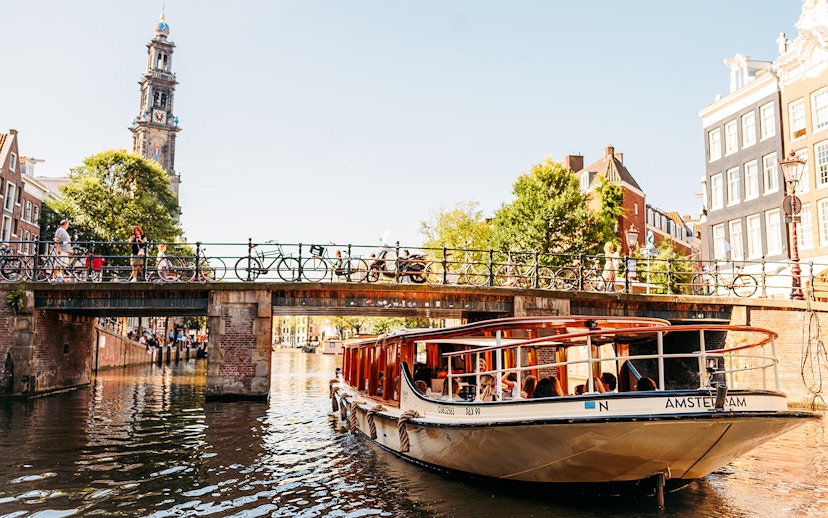Amsterdam canal cruise boat passing under a bridge with Westerkerk tower in the background.