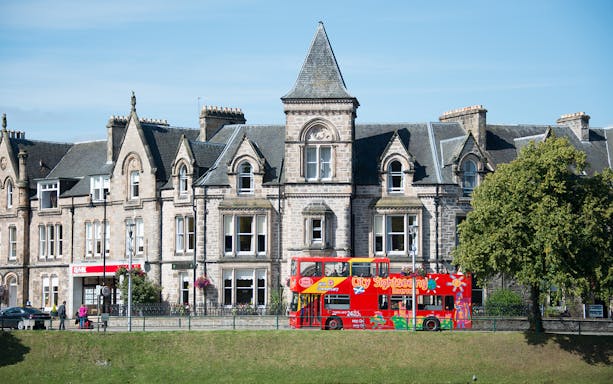 Red double-decker sightseeing bus in front of historic Inverness building on 24-Hr Hop-On Hop-Off Tour.