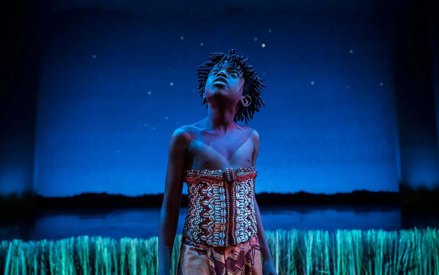 Performer on stage in The Lion King musical, gazing upward under a starry sky.