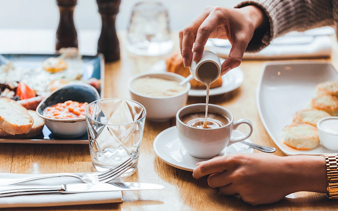 Pouring milk into coffee at breakfast table with pastries.