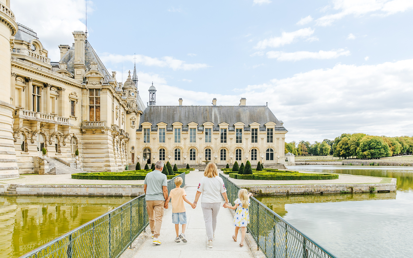 Family walking towards the Chateau of Chantilly, France, across a bridge over a reflective moat.