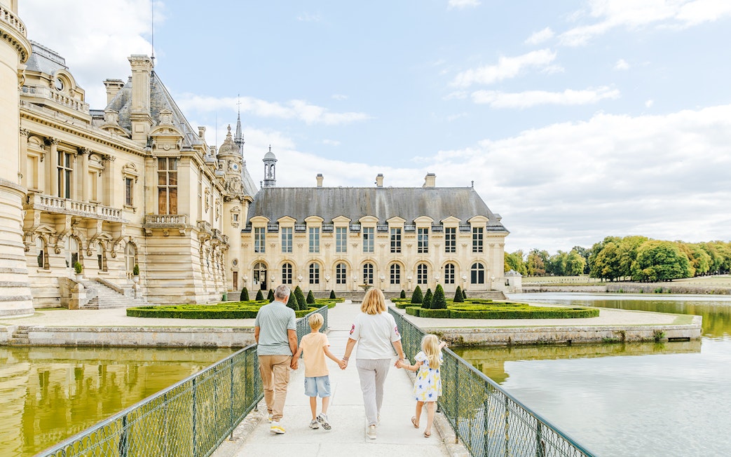 Family walking towards the Chateau of Chantilly, France, across a bridge over a reflective moat.