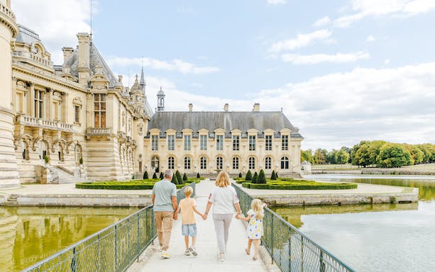 Family walking towards the Chateau of Chantilly, France, across a bridge over a reflective moat.