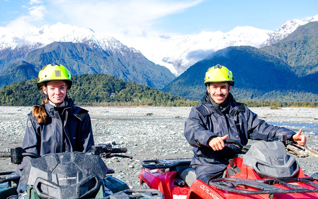 Two people quad biking with snowy mountains at Franz Josef, New Zealand.