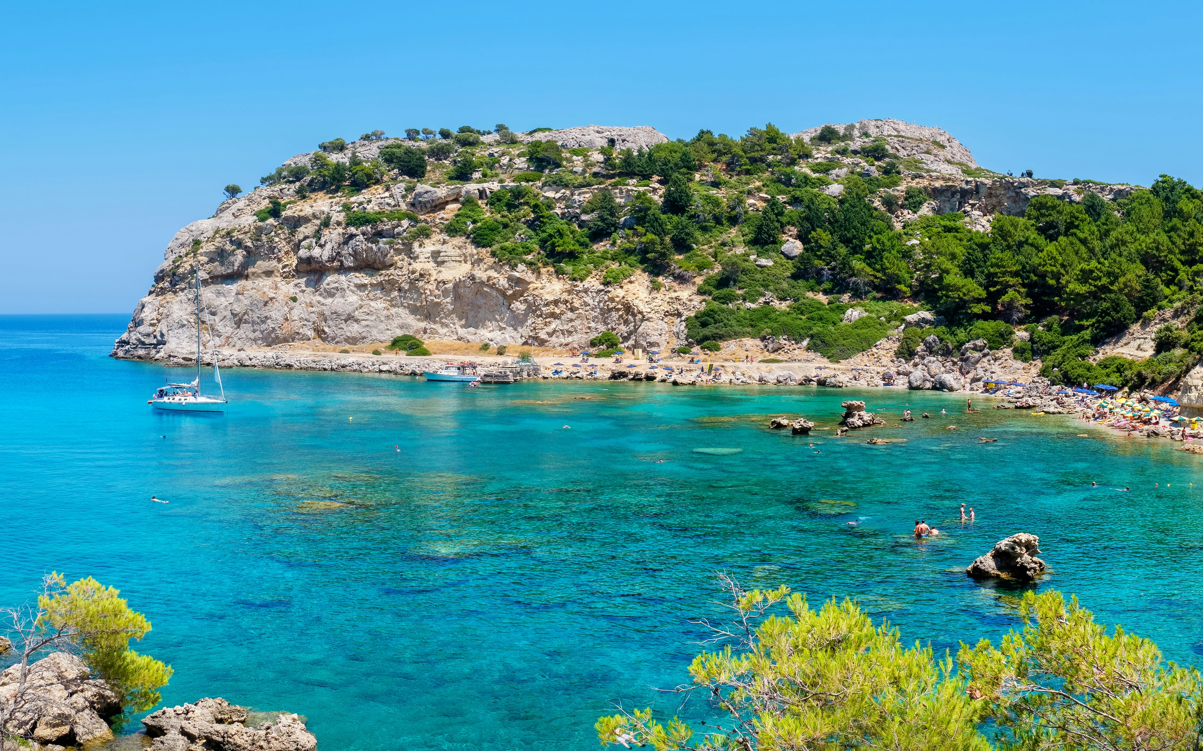 Sailboat in turquoise waters of Anthony Quinn Bay, Rhodes, Greece, with rocky cliffs and beach.