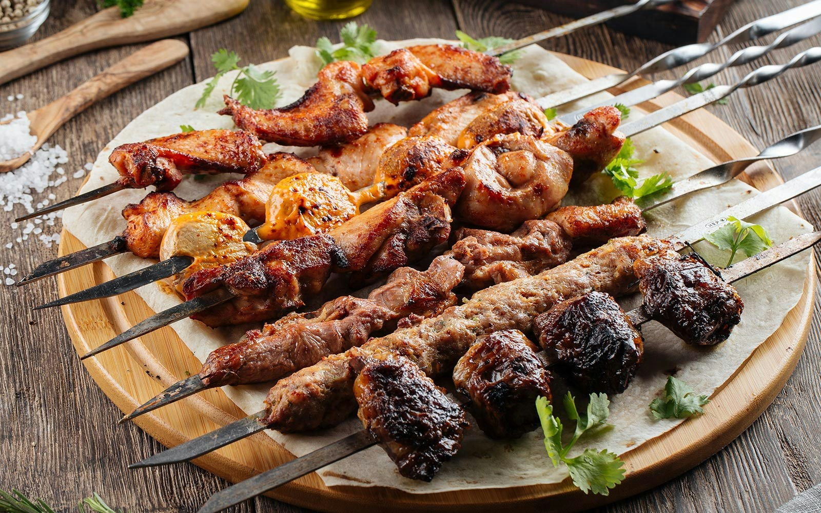 Assorted grilled beef kebabs on skewers with herbs on a wooden platter.