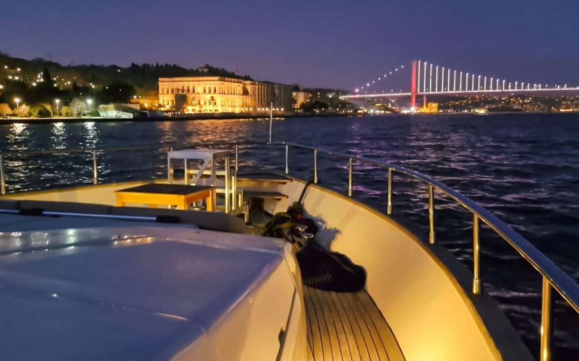 Luxury yacht cruising on Bosphorus at night with illuminated bridge and waterfront buildings.